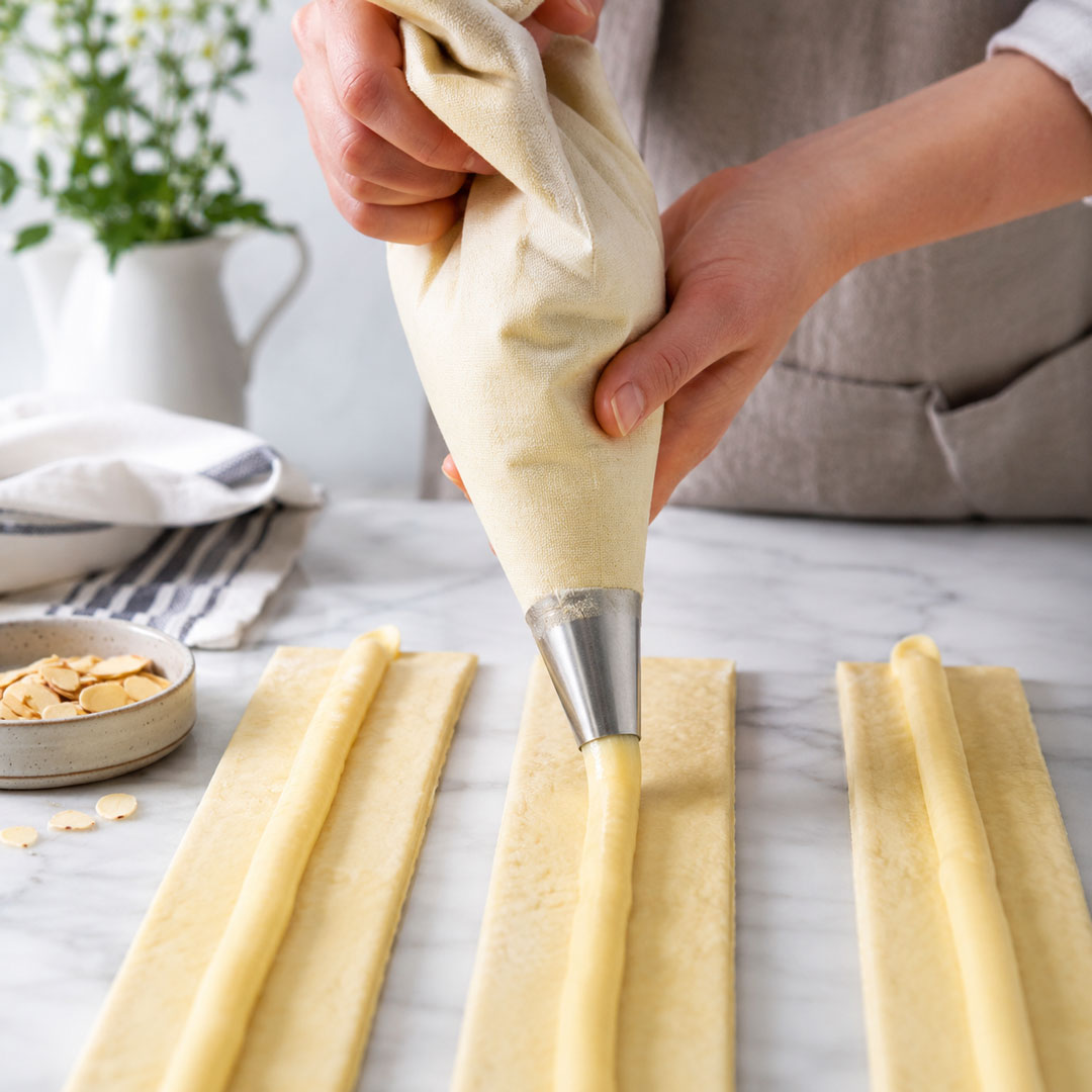 Piping pastry cream onto long strips of dough using a professional piping bag, step-by-step Danish pastry recipe, high-quality food photography.