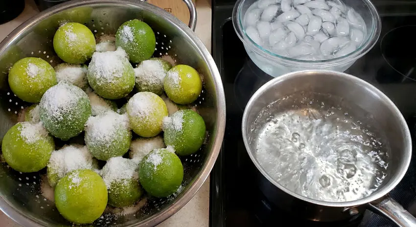 The preparation steps for limes before drying, showing a stainless steel colander filled with small limes heavily coated in salt, a pot of boiling water on a stove, and a large glass bowl containing ice water for a thermal shock bath.